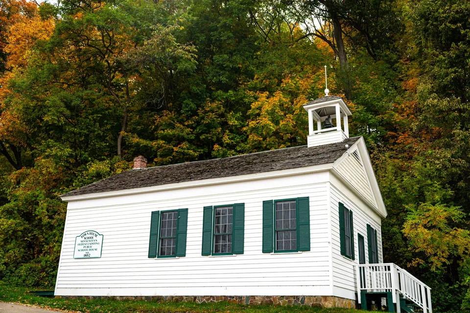 A white historic schoolhouse surrounded by colorful autumn foliage in Taylors Falls, Minnesota.